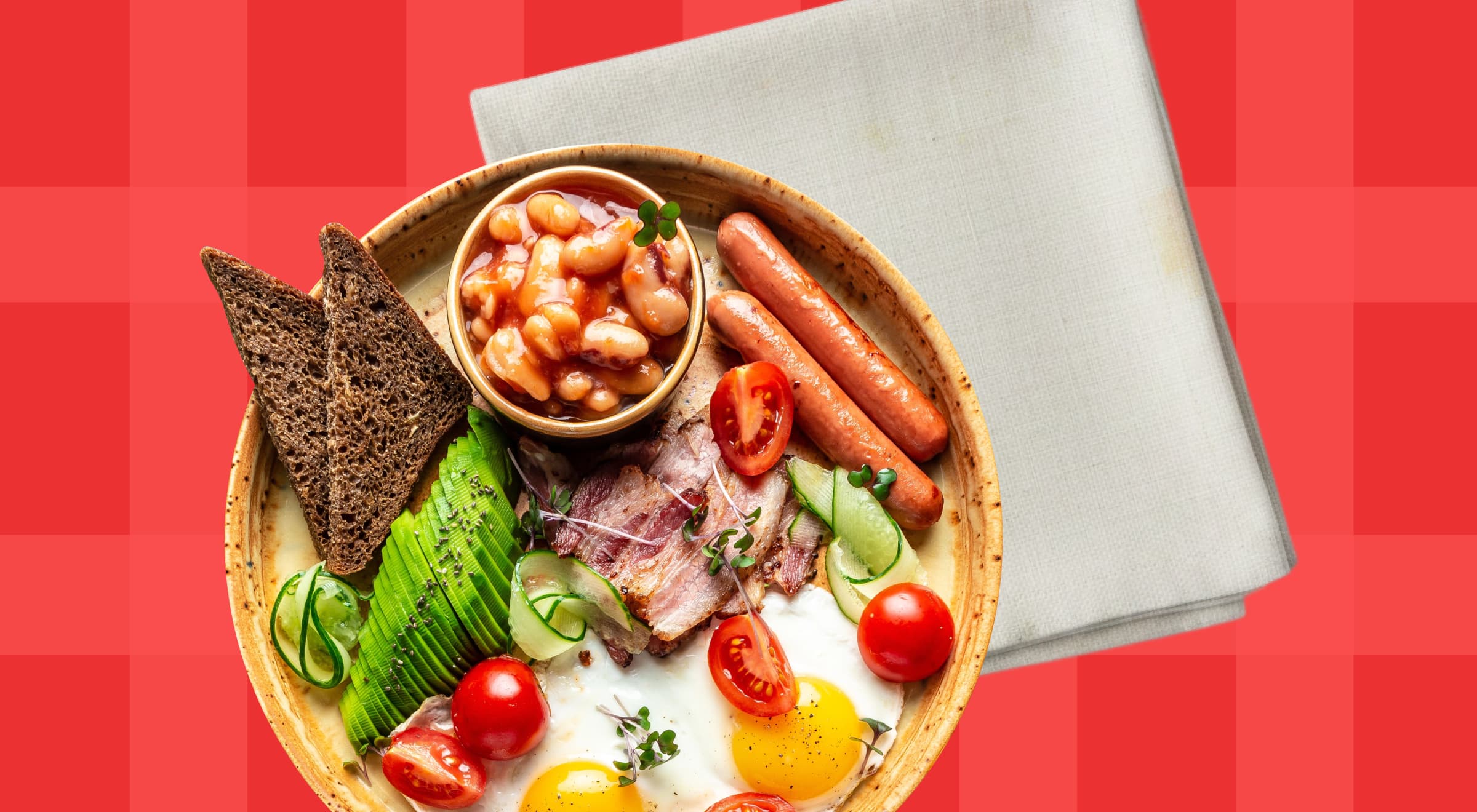 Overhead view of a wooden bowl filled with a full breakfast including fried eggs, sausages, baked beans, cherry tomatoes, dark bread, and greens on a red background