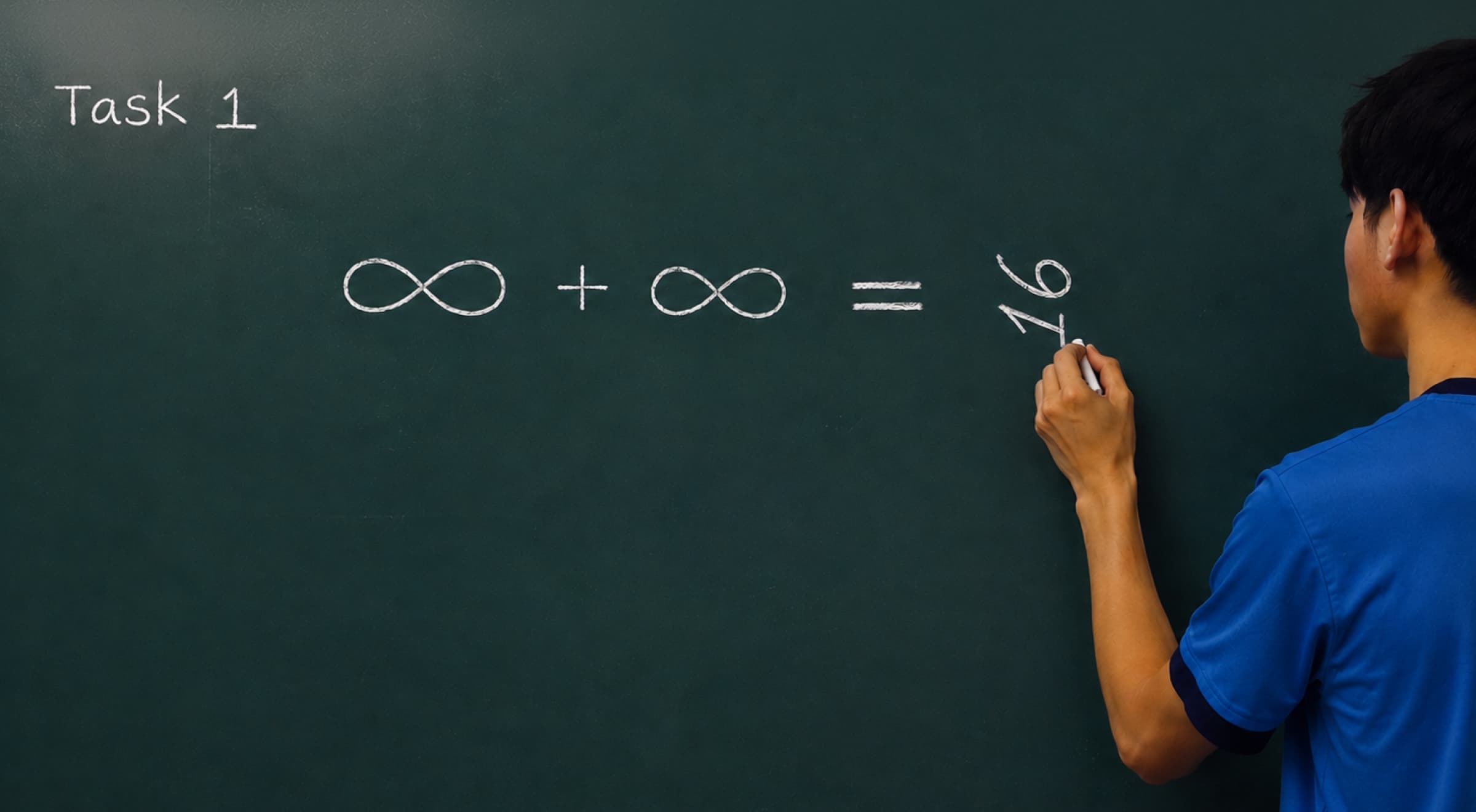 Asian boy in a blue shirt writing a math equation with infinity symbols on a dark green chalkboard in a classroom